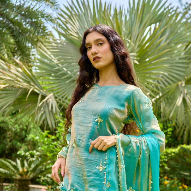 A woman wearing our Mint Green Fendi Silk Garara Set with gold embroidery and a matching dupatta, standing in a lush garden with palm trees.